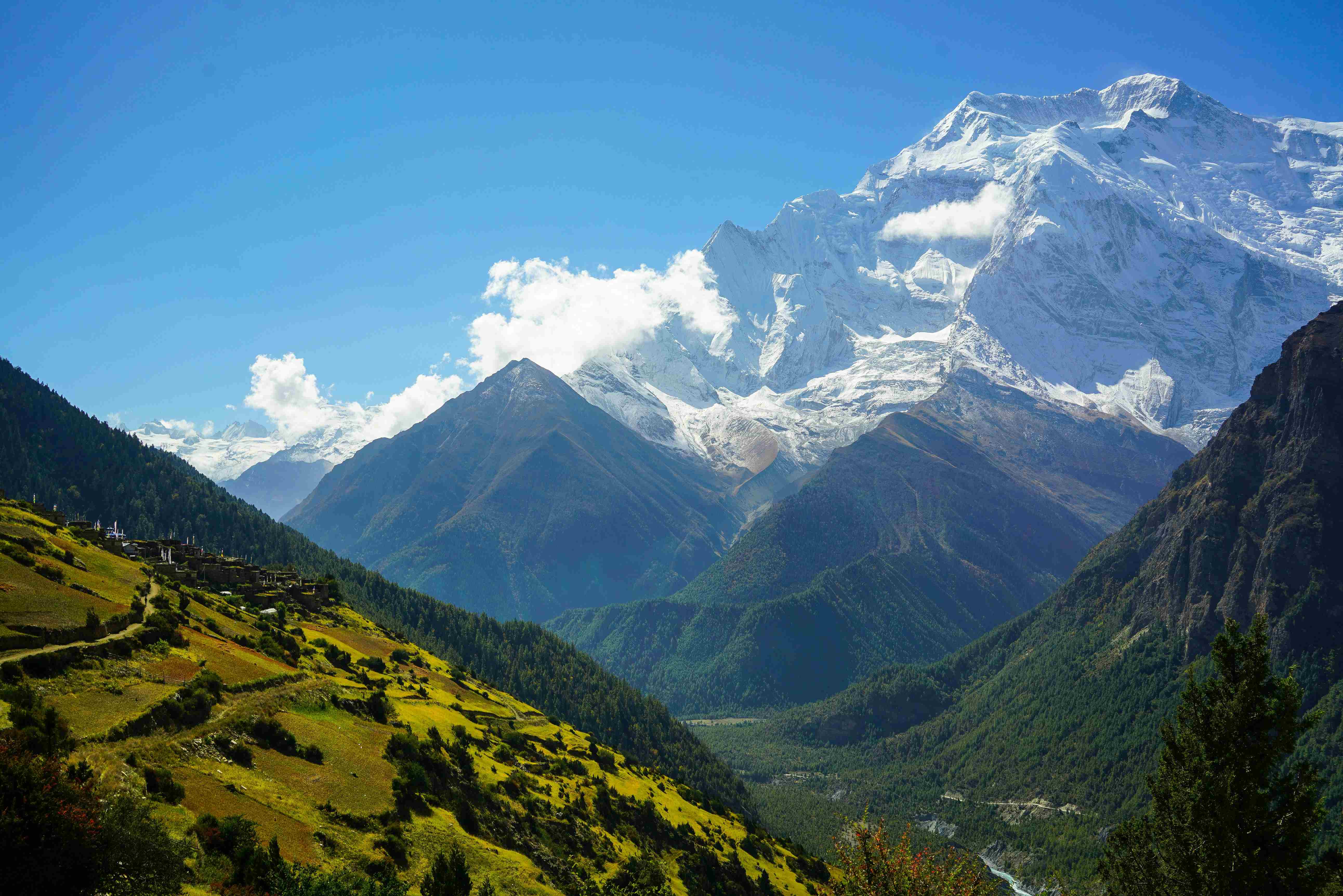A high-angle photograph showing a small village with houses and terraced agricultural fields in a mountain valley, with a large, snow-covered peak towering above.