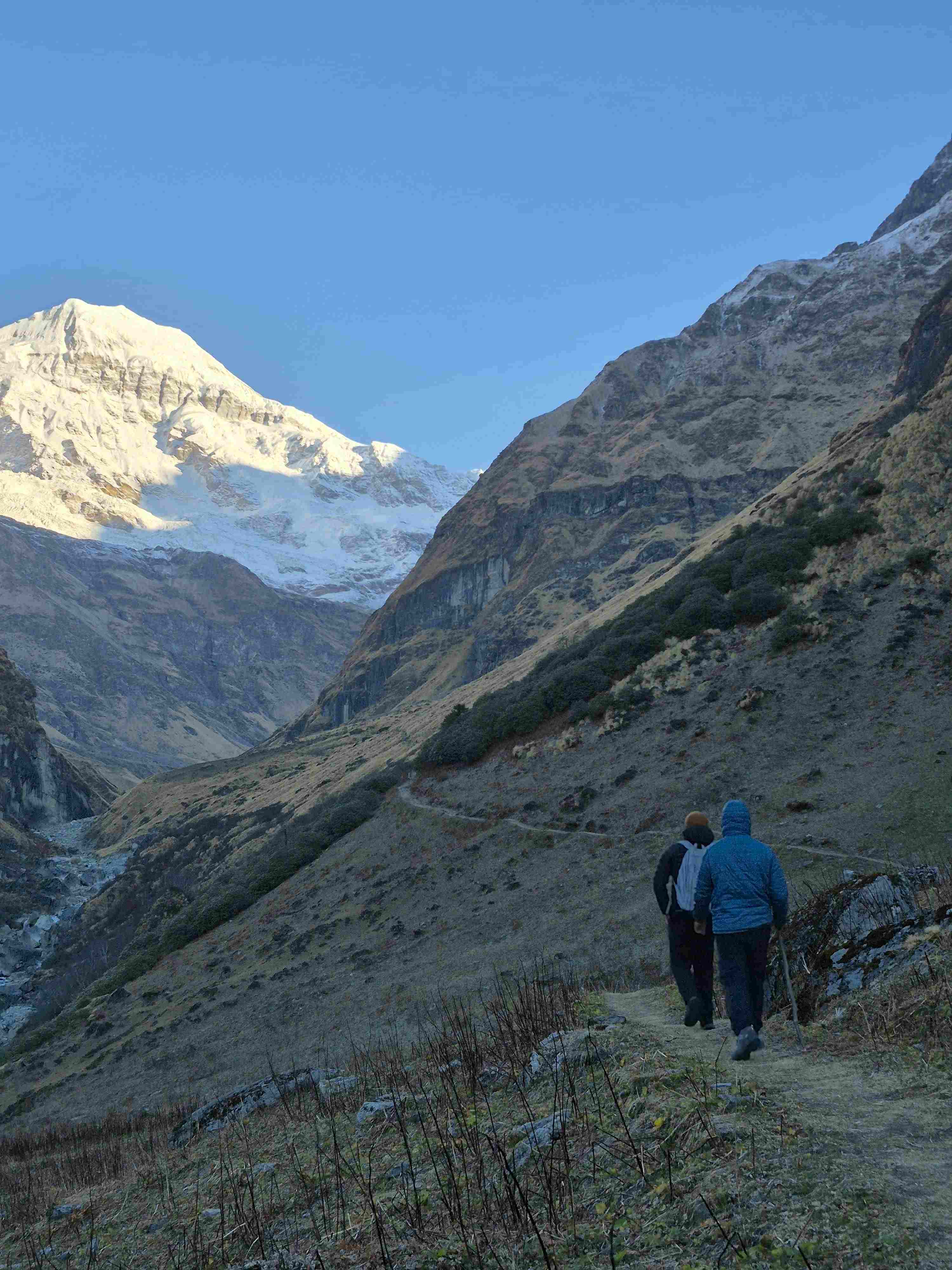 A close-up view of two hikers, with backpacks and walking sticks, on a dirt trekking path in a Himalayan valley with snow-capped mountains in the distance.