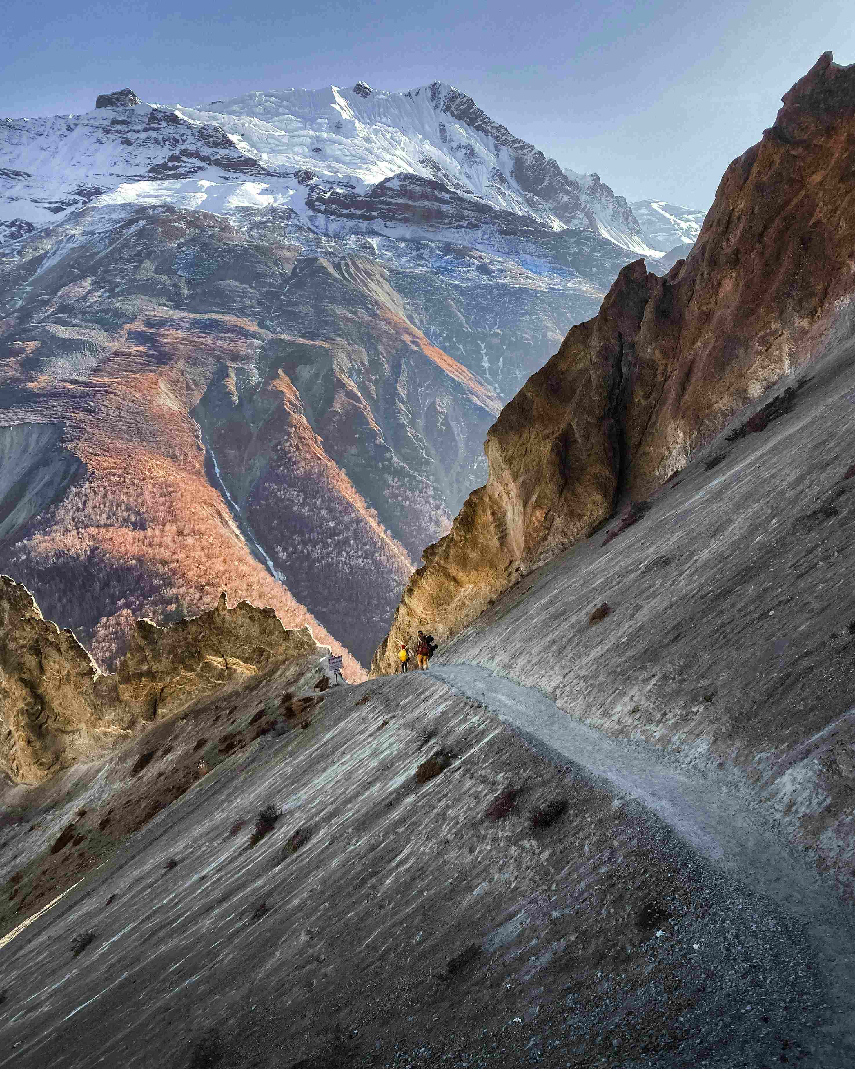 A dramatic photo showing a deep, steep ravine with rocky slopes, and two hikers visible on a narrow trekking path, with a massive snow-capped mountain peak in the background.