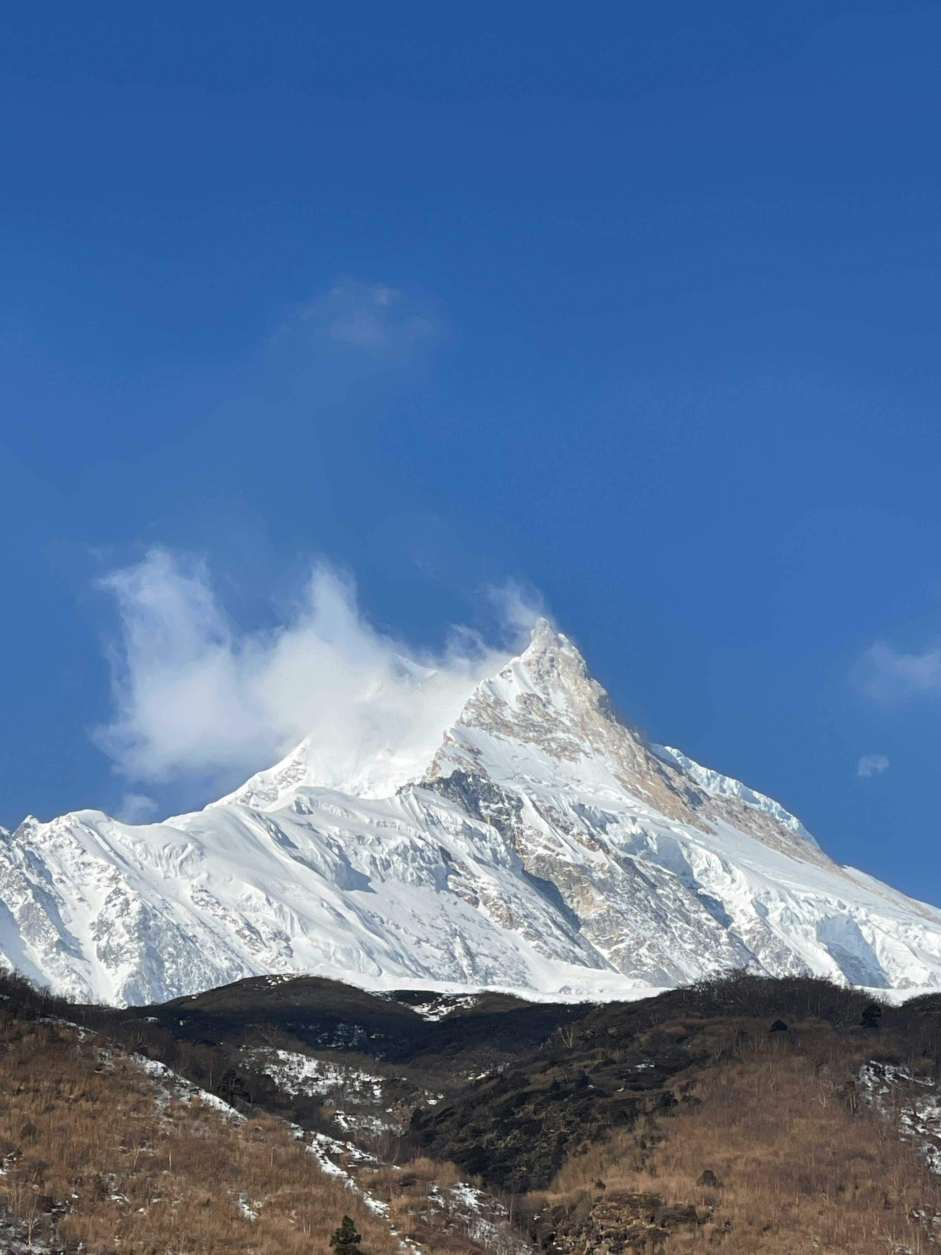 A massive snow-covered Himalayan mountain peak under a clear blue sky with a brown ridge in the foreground 