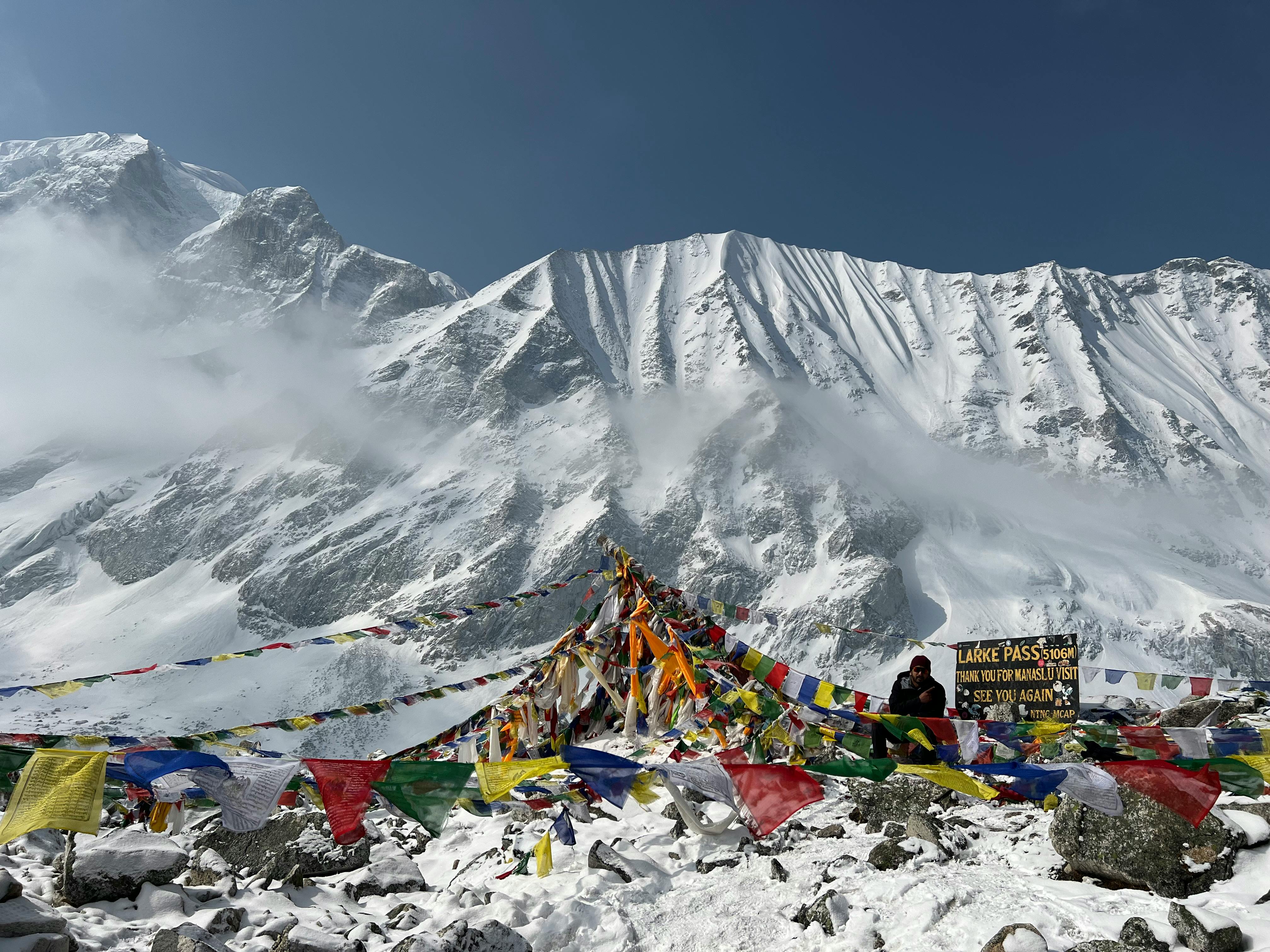 Trekker sitting next to the official Larke Pass summit sign at 5106 meters covered in snow and prayer flags.