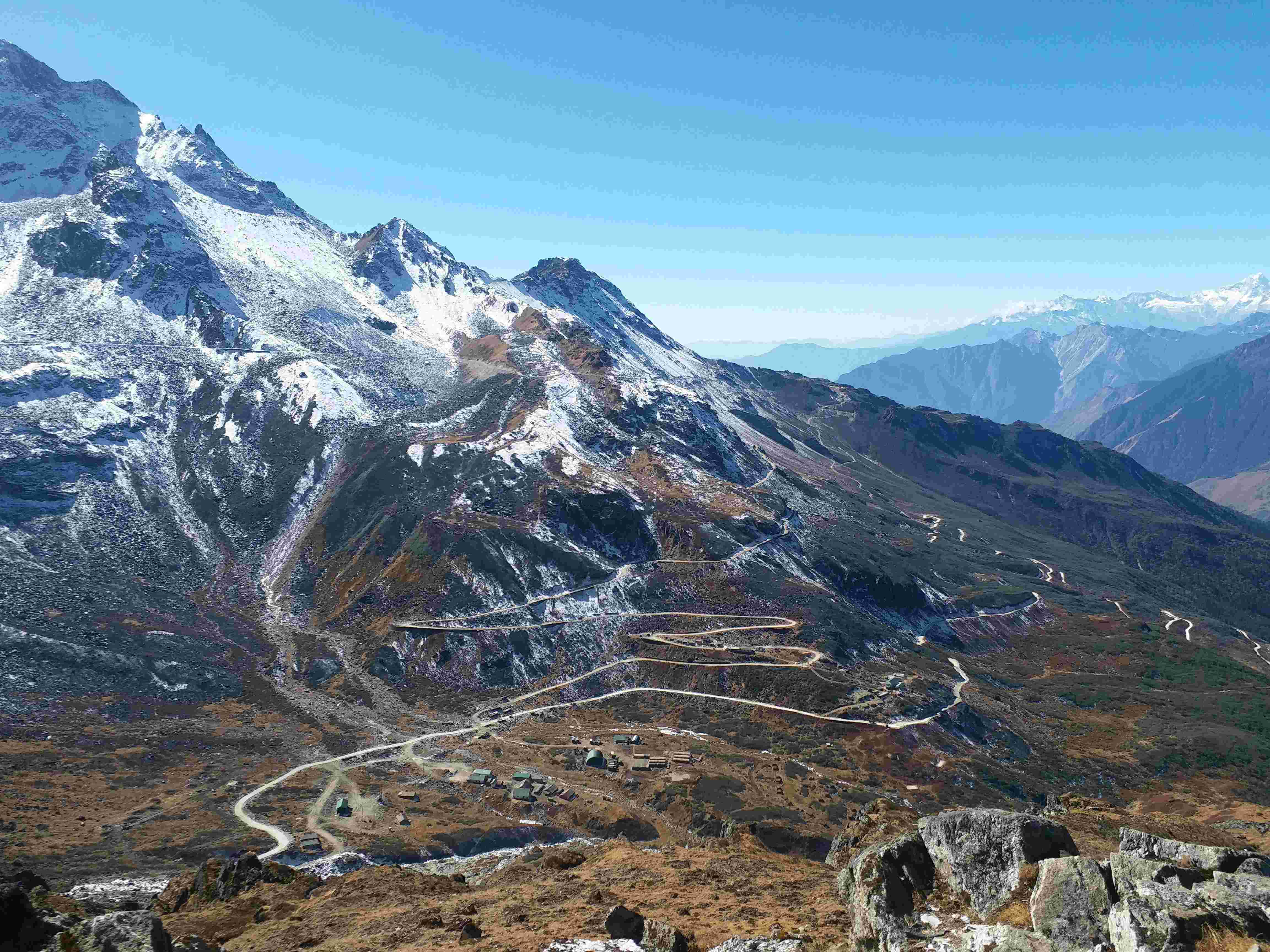 Panoramic view of a steep, zigzagging road carved into the side of a massive mountain with patches of snow and dry vegetation under a bright blue sky.