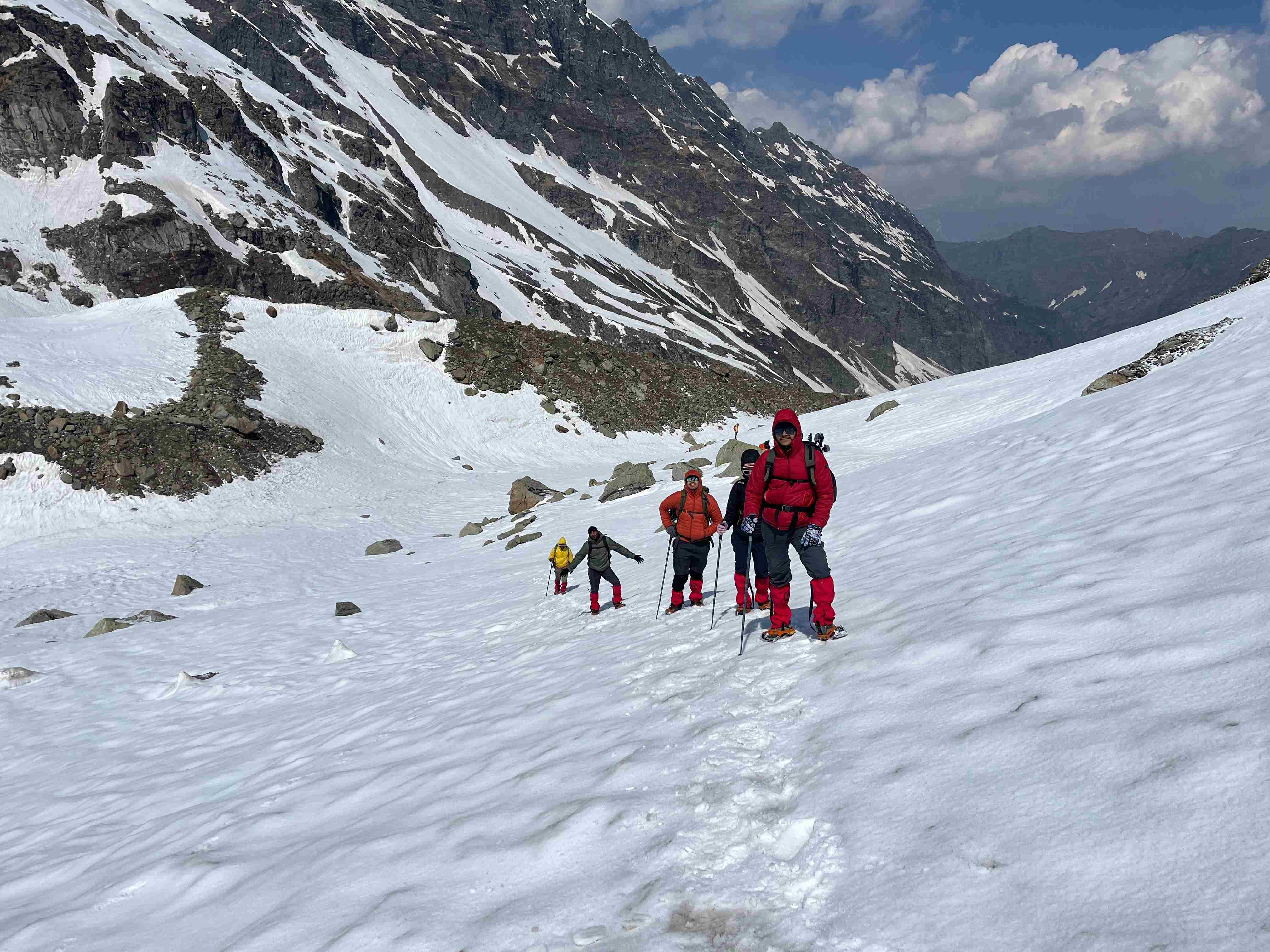 Trekkers in red jackets navigating a steep snow patch during the Hampta Pass trek in the Himalayas.