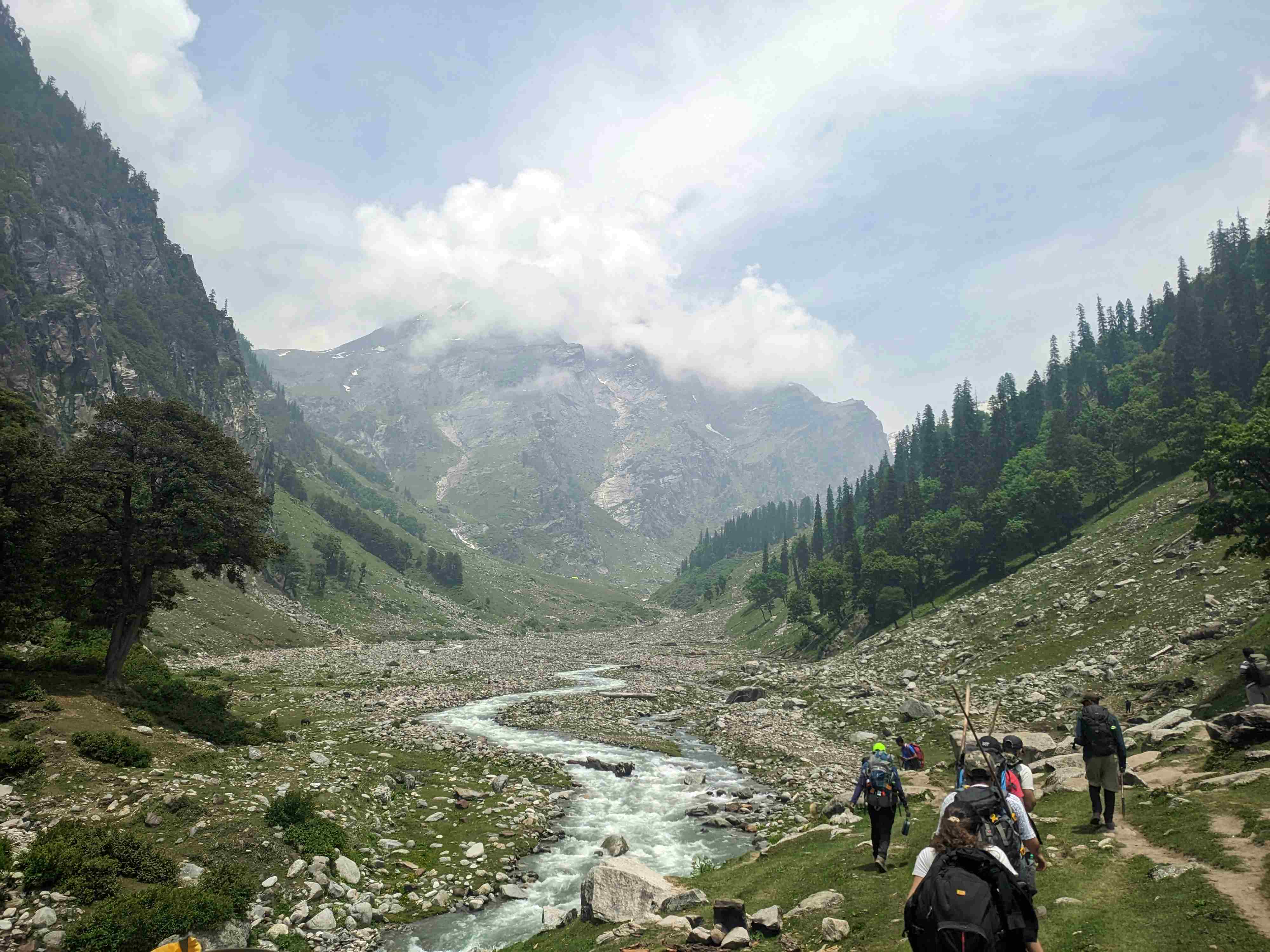 A group of hikers trekking through a lush green Himalayan valley beside a glacial stream.